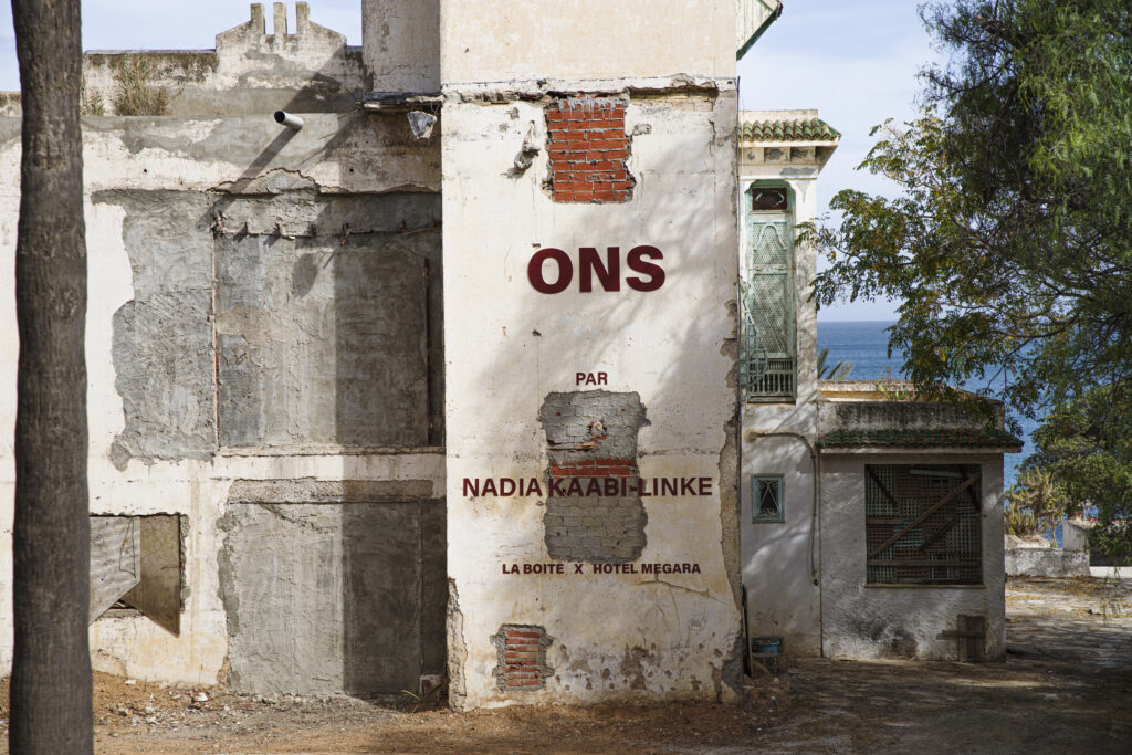 On the facade of an abandoned building, the words ONS are written in large letters, with further text underneath. In the background, you can see the sea. Nadia Kaabi-Linke Kaf w Kaaba Halwa