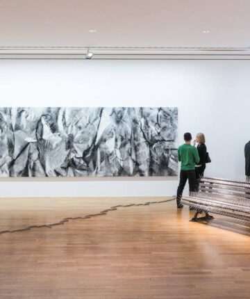 Visitors standing in between artworks in an exhibition space. A park bench covered with bird spikes, a crooked porcelain line on the ground and a big canvas on the wall. Nadia Kaabi-Linke, Versiegelte Zeit.