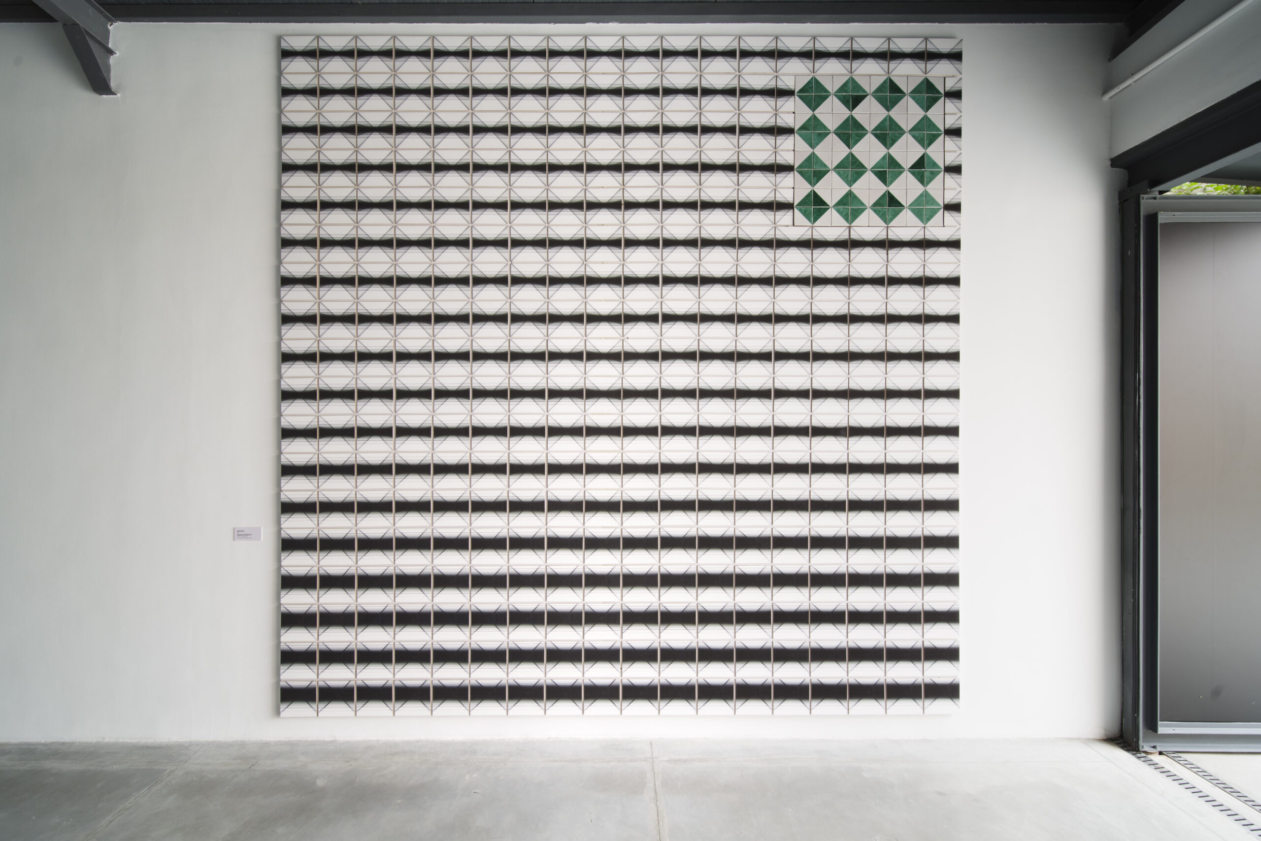 An artwork made of many pictures aligned in a big square, creating a black and white pattern, in the right corner some pictures create another pattern in green, hanging on the wall of an exhibition space