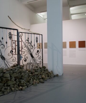 A cast-iron gate with adjacent stones separates an exhibition space. In the background rectangular pictures of different colors, hang on the wall.