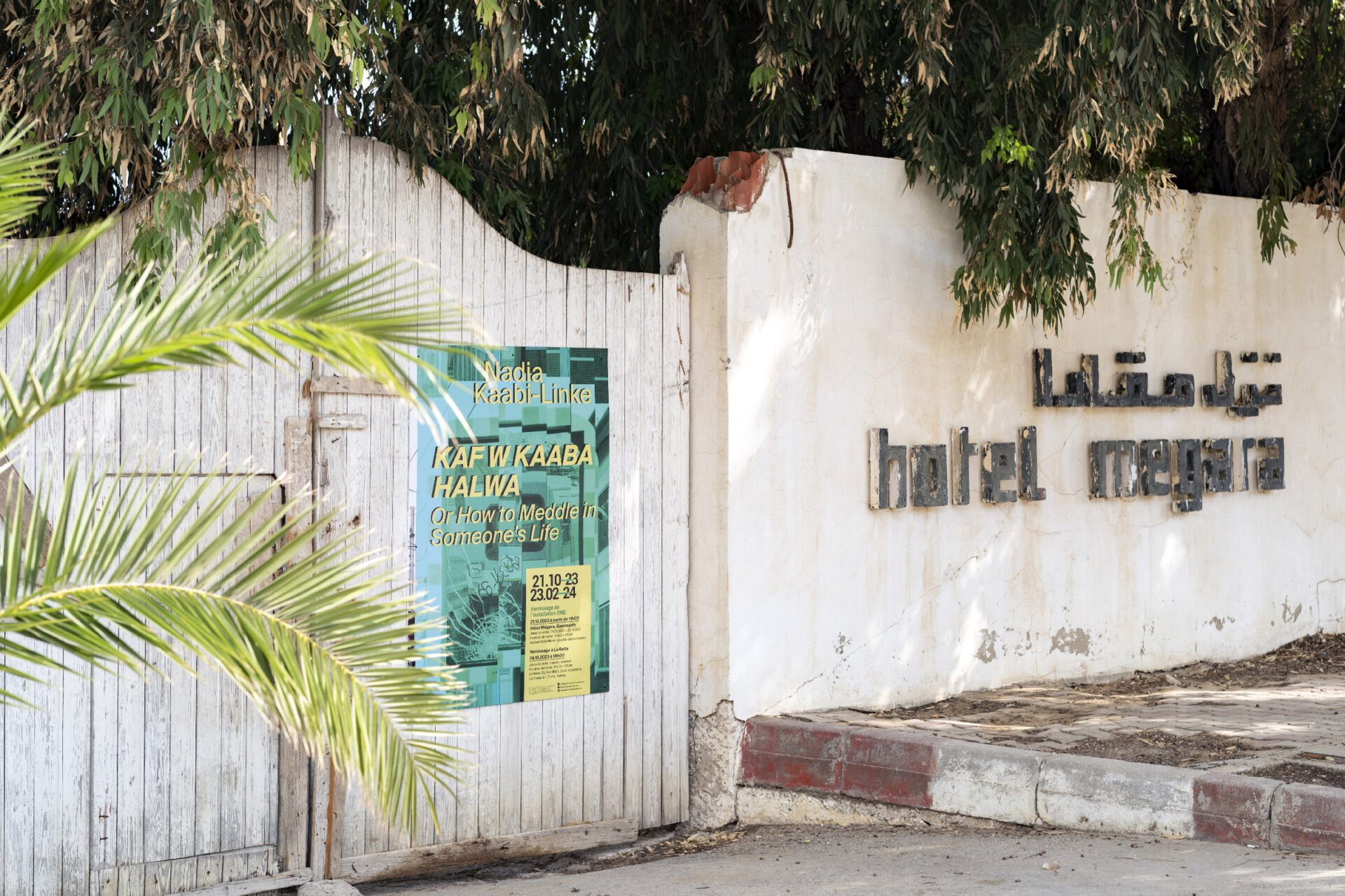A white, wooden gate, that was once the entrance to the Hotel Megara Gammarth, is now holding the poster about an exhibition.