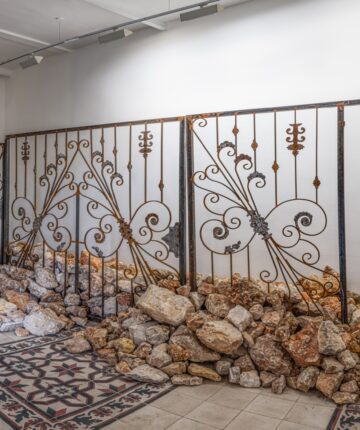 In an exhibition space a Detail of a cast iron gate is standing in between a pile of stones.