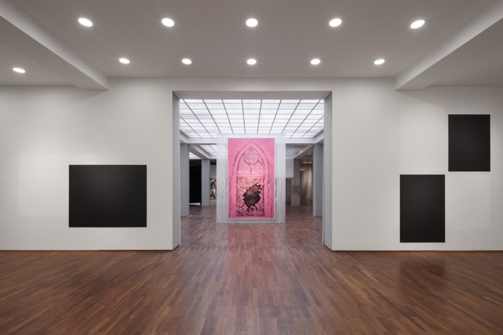 Black squares hanging on the walls of an exhibition space, in the background, another room, with a big pink painting.