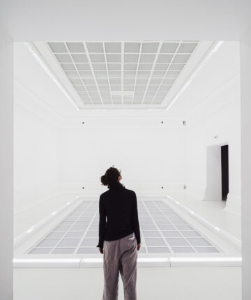 A woman photographed from behind, standing in a large white exhibition room, a large floor construction reflected on the ceiling. Nadia Kaabi-Linke, Heaven and Hell.