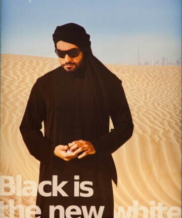 A man stands in the desert, with Dubai in the background. The title reads "Black is the new white."
