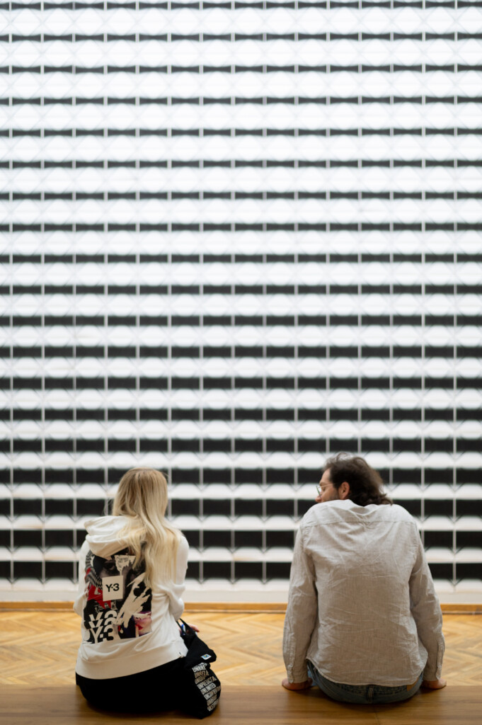 Two people sitting in front of an artwork, photographed from behind.