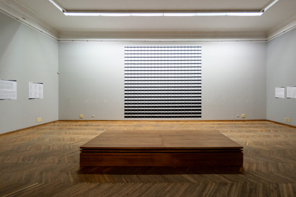 An exhibition space with a big wooden bench in the center. On the wall a big artwork consisting of many little images