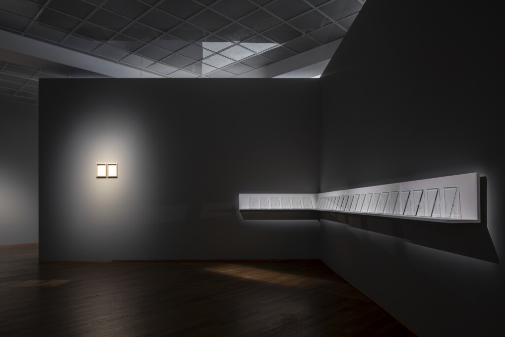 The corner of an exhibition space. A row of glass squares sitting on a shelf and two little frames are illuminated.
