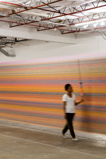 A person walks along two columns holding a vibrant, multicolored thread, showcasing a blend of orange, pink, and blue hues. Nadia Kaabi-Linke, "Walk the Line".