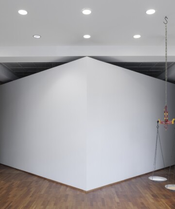 An exhibition space featuring a large, angular white structure positioned centrally, creating two narrow passageways on either side. A red and yellow industrial lifting device hangs from the ceiling, suspended a scale one part salt one part sand. Recessed ceiling lights evenly illuminate the minimalist room, and a glimpse of another installation with vertical hanging strings is visible through the left passage. Nadia Kaabi-Linke, Die Alte Mauer.