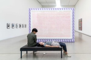 View of an Exhibition space with a big canvas in the middle of the room hiding a big part of it behind it. In front of the canvas there is a black bench with a boy sitting on it and next to him a girl laying on its back, foots on the ground.