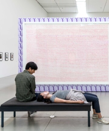 View of an Exhibition space with a big canvas in the middle of the room hiding a big part of it behind it. In front of the canvas there is a black bench with a boy sitting on it and next to him a girl laying on its back, foots on the ground. Nadia Kaabi-Linke, Nervous Bench.