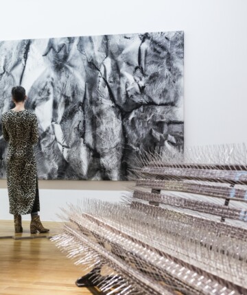 in the front of the image, a park bench covered with birth spikes. Behind it a visitor of the exhibition space is looking at a big black and white canvas on the wall.