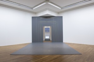 Geometric art installation with a gray panel, rectangular gold outlines, and a doorway in the center, positioned in a white-walled gallery room with wooden flooring.
