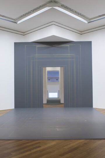 Geometric art installation with a gray panel, rectangular gold outlines, and a doorway in the center, positioned in a white-walled gallery room with wooden flooring. Nadia Kaabi-Linke, Versiegelte Zeit.