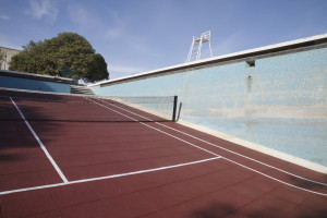 A red tennis court with a net, set in an empty swimming pool and a lifeguard chair, surrounded by greenery and a clear blue sky.