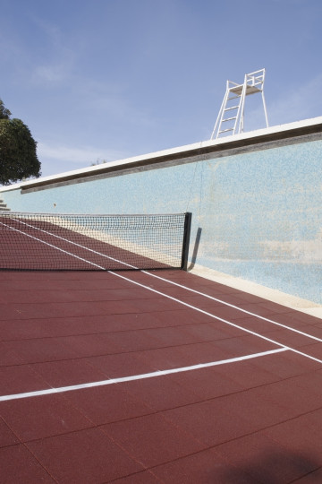 A red tennis court with a net, set in an empty swimming pool and a lifeguard chair, surrounded by greenery and a clear blue sky. Nadia Kaabi-Linke ONS