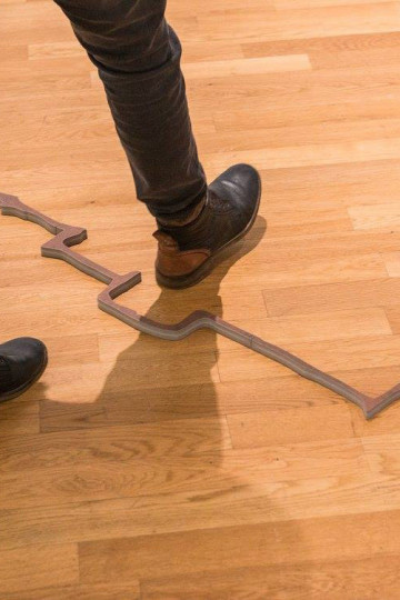 Someone, only visible from the knees down, stepping over an uneven brown line laying on a wooden floor. Nadia Kaabi-Linke, Gekochte Erde.