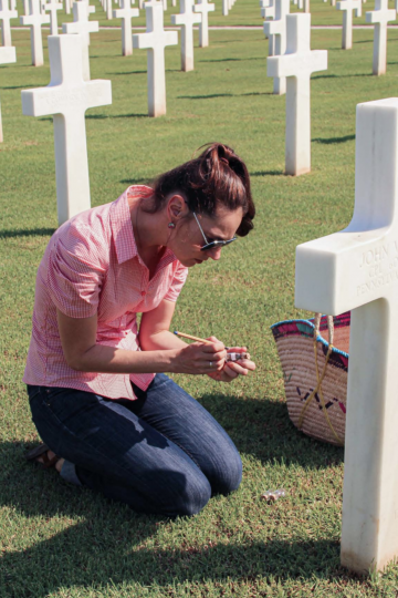 The artist Nadia Kaabi-Linke sits infront of a gravestone, a white cross, in the North Africa American Cemetery and Memorial in Tunisia, to copy the engravings onto a small flacon/ Tunisian Americans, 2012. Artwork by Nadia Kaabi-Linke