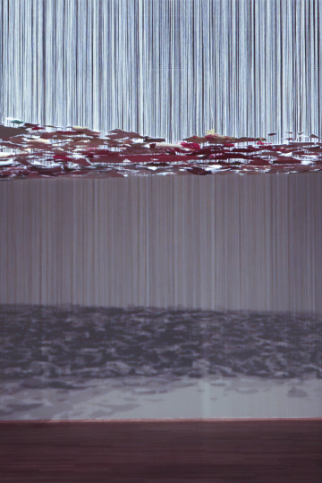 Colored wall pieces with paint hanging on white threads are casting shadows on the museum's wall. Nadia Kaabi-Linke, Under Standing Over Views.