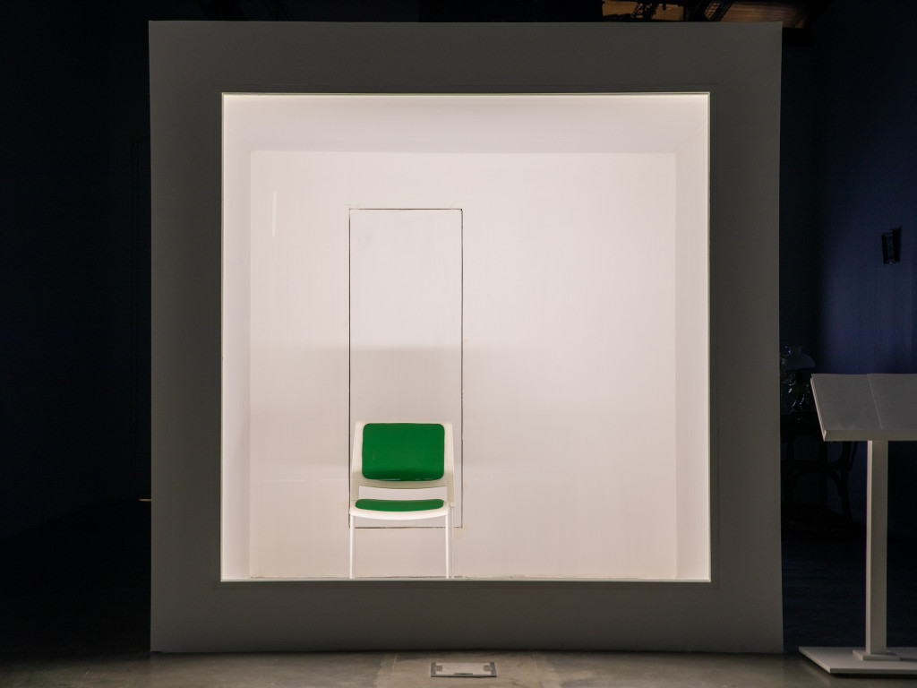 A white box placed in a dark room. Inside the heavily lit Box, a white chair with green padding.