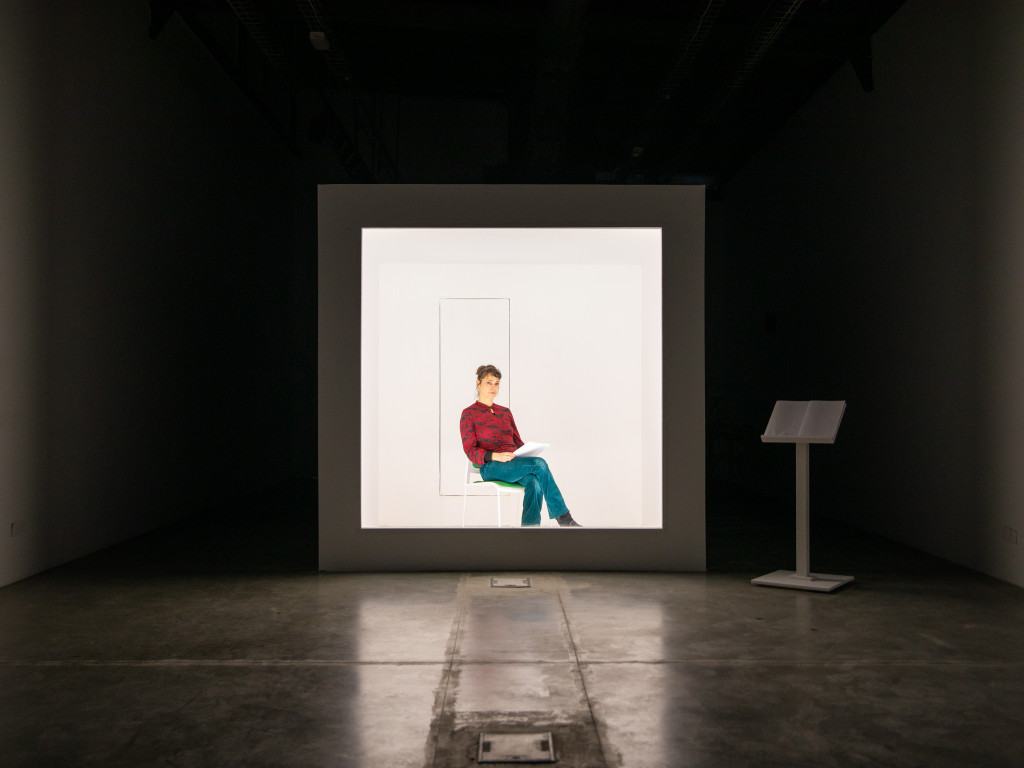 A white box placed in a dark room. Inside the heavily lit Box, the artist Nadia Kabbi-Linke sitting on a chair holding a bunch of papers.