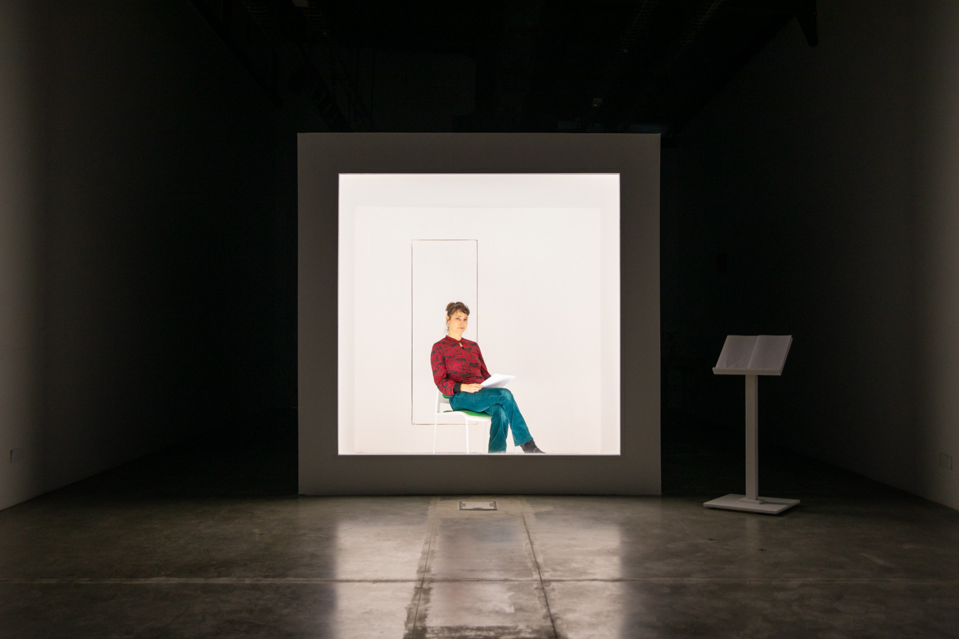 A white box placed in a dark room. Inside the heavily lit Box, the artist Nadia Kabbi-Linke sitting on a chair holding a bunch of papers.