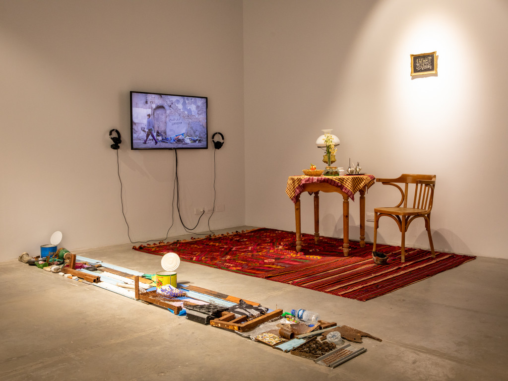Underneath a framed image a set table with a wooden chair on a colorfull carpet. Next to it, on the wall, a TV showing a video of a woman looking for objects on the floor in front of a destroyed wall. On each side of the screen, a headphone. In front of the carpet, a line of found objects.
