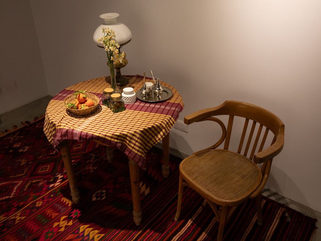 A wooden table covered by a tablecloth. On the table, a lamp, flowers, tea and a bowl with nectarines. next to it a wooden chair. The floor is covered by a colorful carpet.