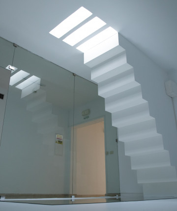 A white room with an upside down staircase which ends on a mirror laying on the ground. On the ceiling, the staircase continues in the form of lights in the same size of the steps. On the left side, a glass wall gives view into a hallway.