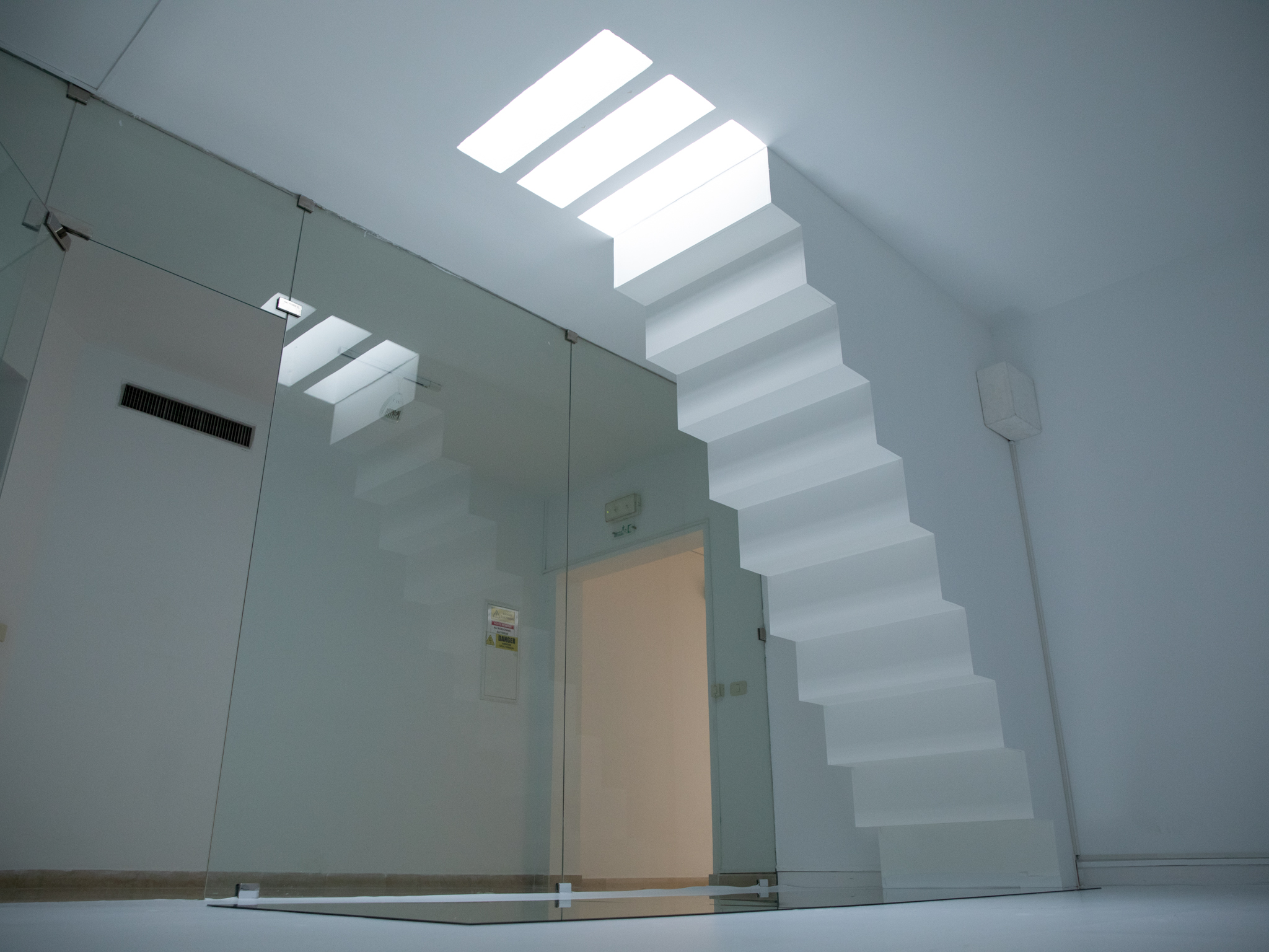 A white room with an upside down staircase which ends on a mirror laying on the ground. On the ceiling, the staircase continues in the form of lights in the same size of the steps. On the left side, a glass wall gives view into a hallway.