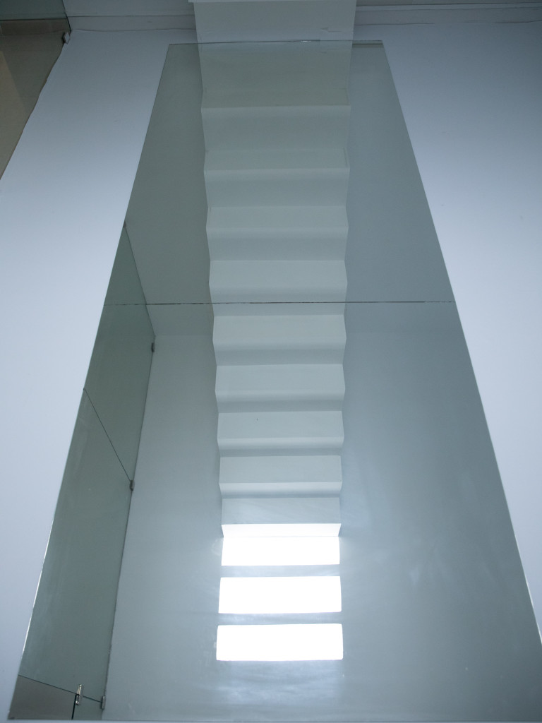 A large mirror on the ground reflecting a white staircase and three cubes of light in the same size of the steps.