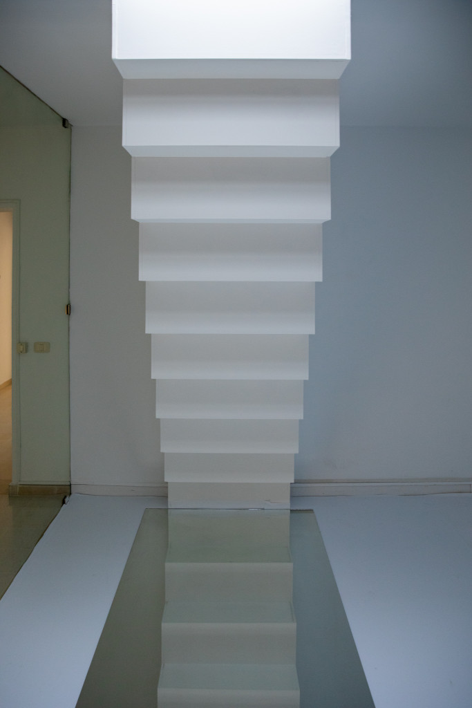 A white room with an upside down staircase which ends on a mirror laying on the ground. The room is illuminated from the ceiling. On the left side, a glass wall gives view into a hallway.