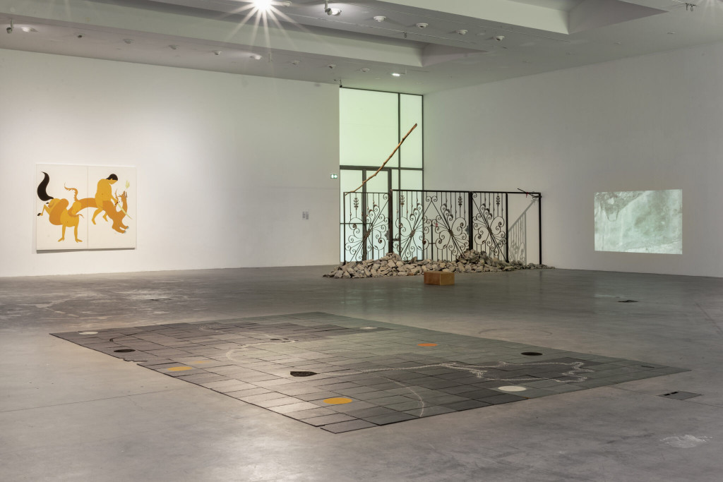 Exhibition space with different artworks scattered around. On the wall a painting with mystical figures, on the ground an installation with black tiles. In the back a cast iron gate with stones and branches. A video on the wall next to it.