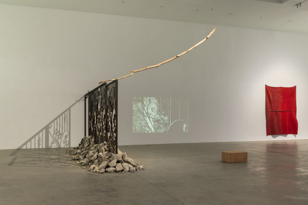 Exhibition space with an installation consisting of an iron cast gate, stone rubbles, branch and video. A red fabric is hanging on the wall.