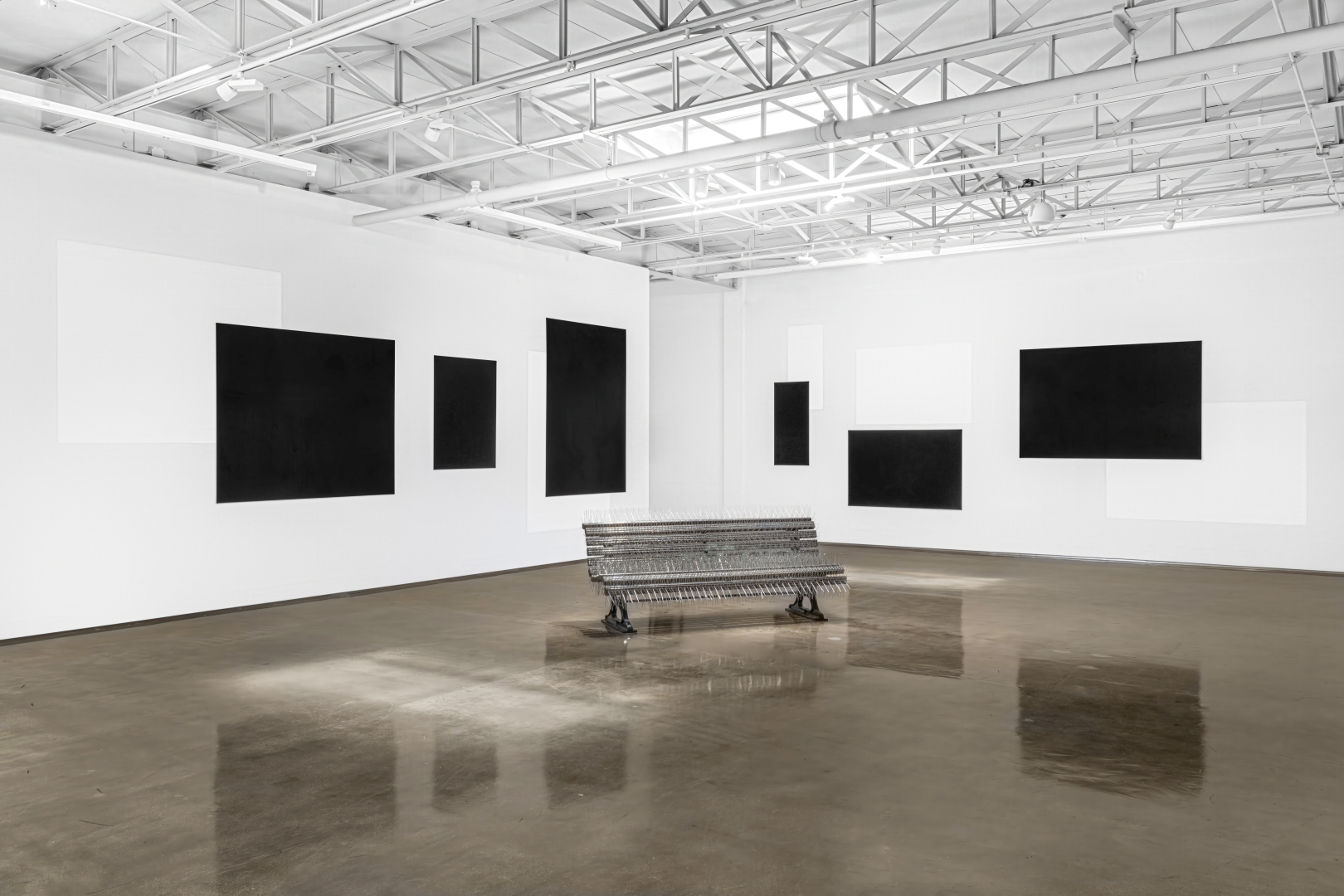 White gallery space with an industrial ceiling and a spiked park bench, black panels, and white shapes on a white wall.