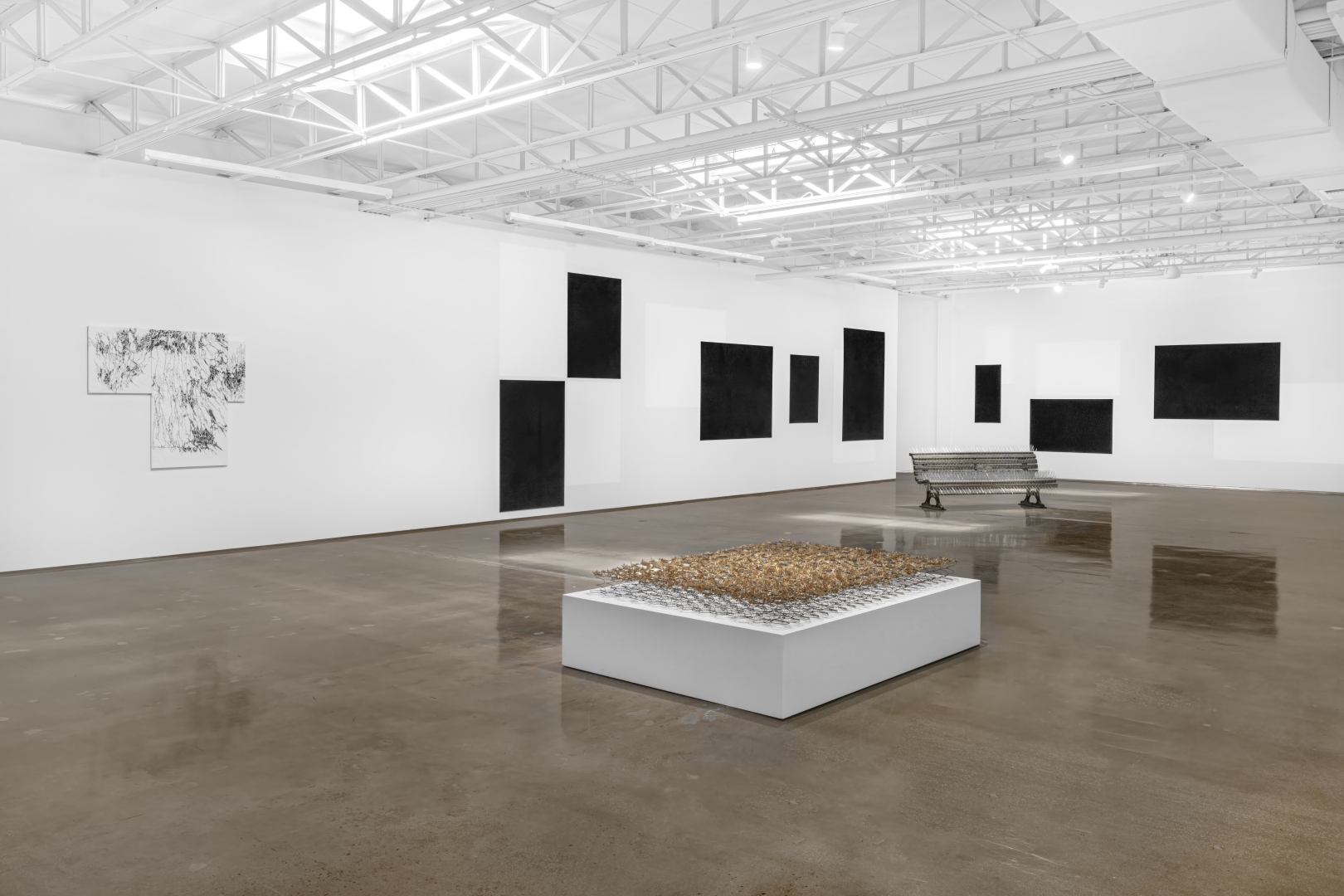 White gallery space with an industrial ceiling and sculpture with dried thistles, black and white drawings on three canvasses, a spiked park bench, black panels, and white shapes on a white wall.
