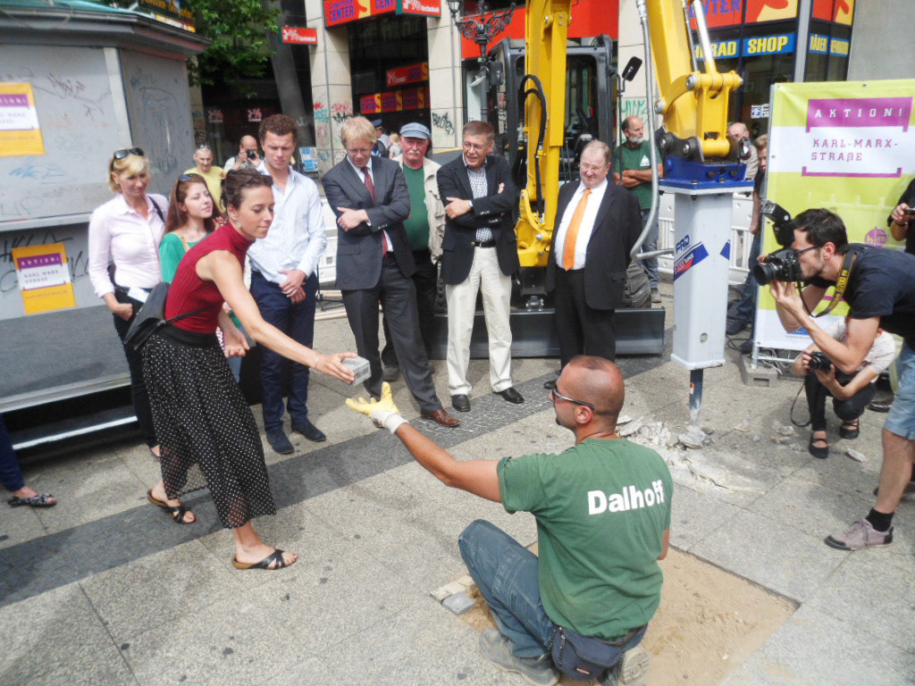 A group of people standing around a hole in the ground, where a man is adding a paving stone handed to him by a bystanding woman.