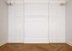 View of an exhibition space, whit walls and wooden floor. The wall in the back has thin brass lines that continue on the floor, creating different sized squares.