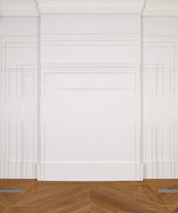 View of an exhibition space, whit walls and wooden floor. The wall in the back has thin brass lines that continue on the floor, creating different sized squares. Nadia Kaabi-Linke, Modulor II.