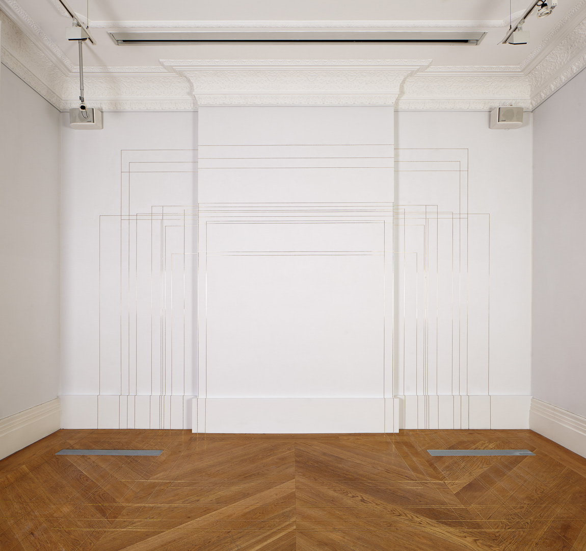 View of an exhibition space, whit walls and wooden floor. The wall in the back has thin brass lines that continue on the floor, creating different sized squares.