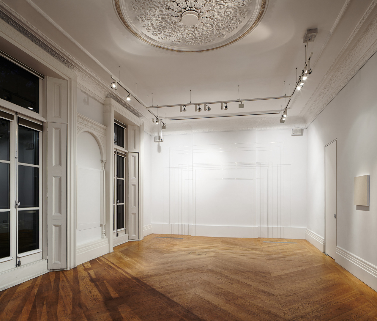 View of an exhibition space, whit walls and wooden floor. The wall in the back has thin brass lines on it.