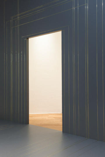 A grey room with metal inlays, a opening in the middle of the image gives way into another brighter room.
