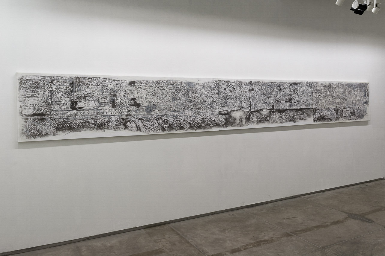 A long canvas with frottage pattern, taken from a wall with cracks and engraved names, hanging on an exhibition wall.