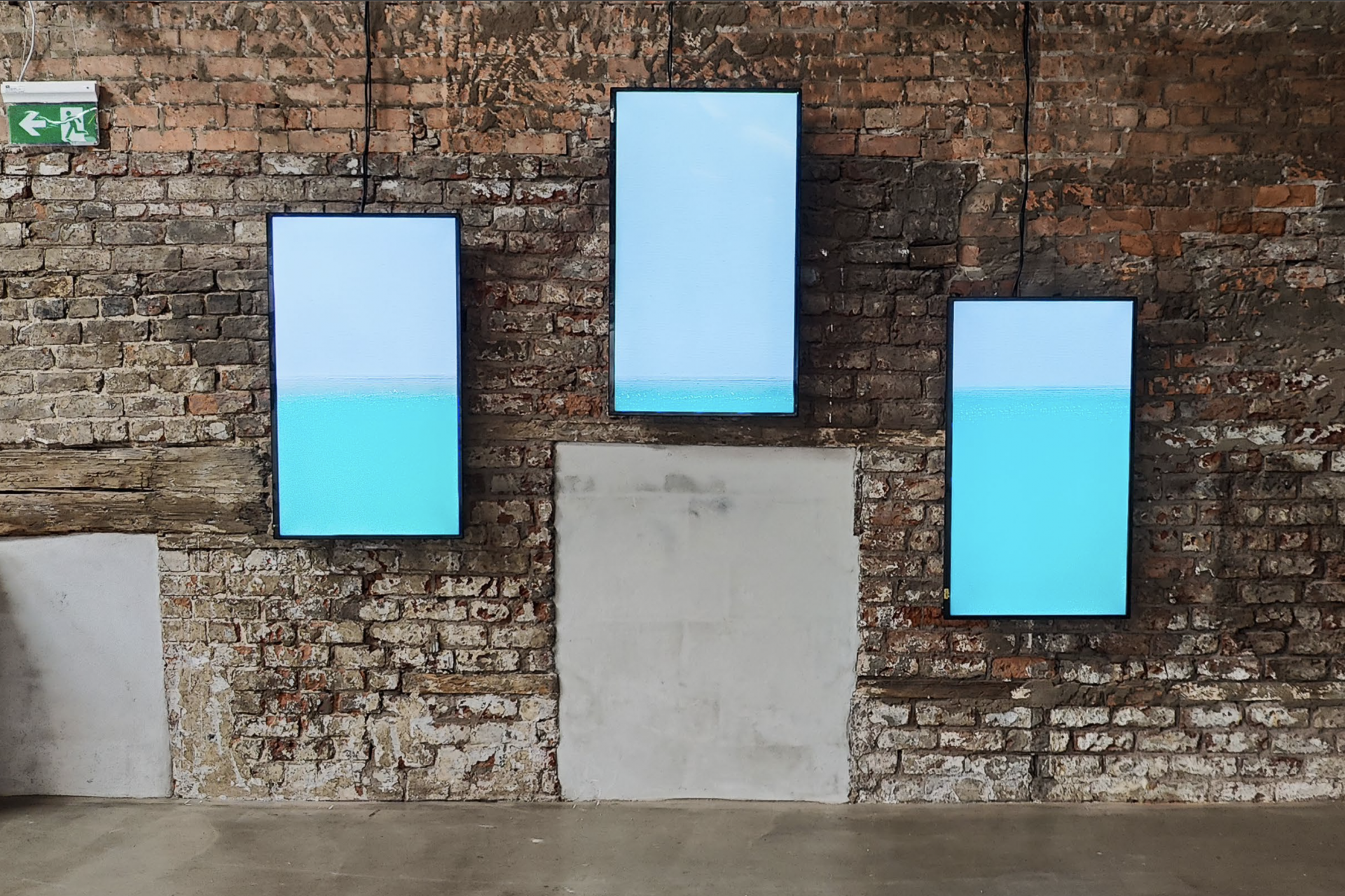 A video of the sea divided on three vertical screens hanging on a brick stone wall. Nadia Kaabi-Linke Under Sea Heart Beats