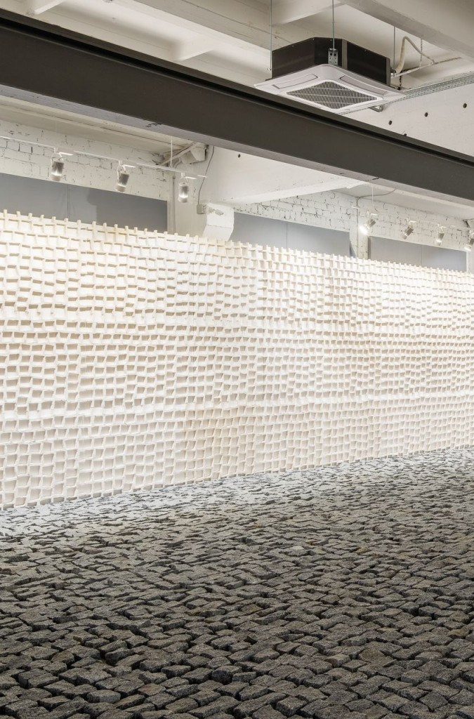 Installation view of a white wall with holes in the form of cobblestones. On the ground in front, the cobblestones.