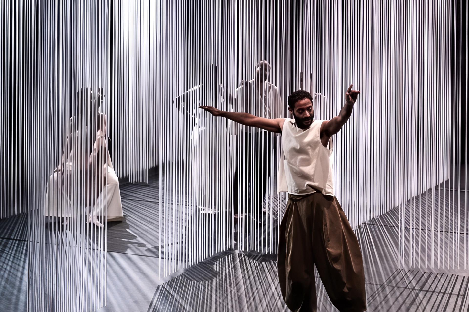 A man is dancing in front of white string curtains, while four other performers behind the curtains are watching him.