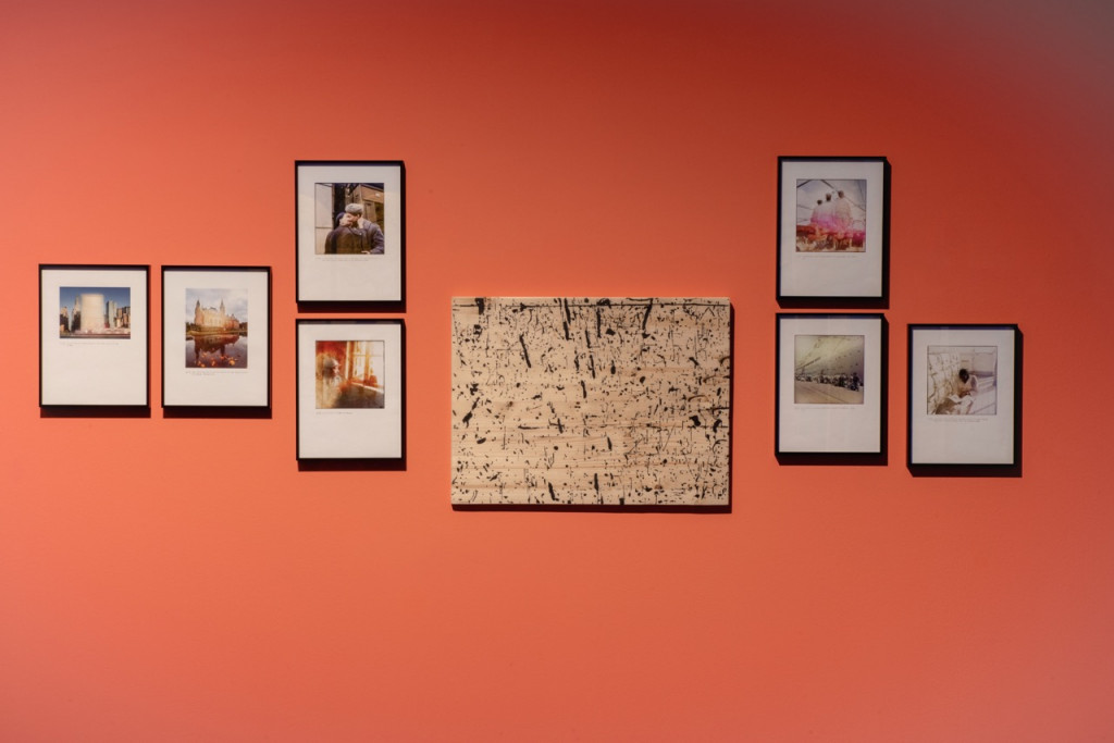 Photographs in black frames and a wood panel with black marks on an orange wall.