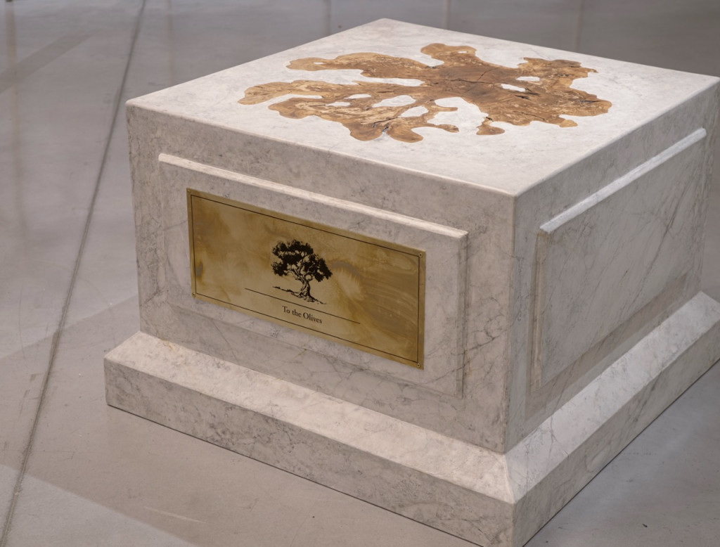 Nadia Kaabi-Linke, To the Olives, 2026. Marble plinth sculpture with olive trunk cross-section on top and brass plaque engraved with an olive tree, seen on a grey floor.