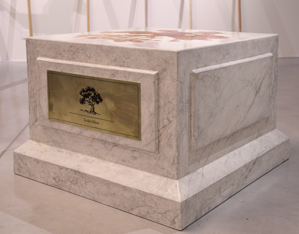 Nadia Kaabi-Linke, To the Olives, 2026. Marble plinth sculpture from a low angle, showing the brass olive tree label, framed panels and stepped base.